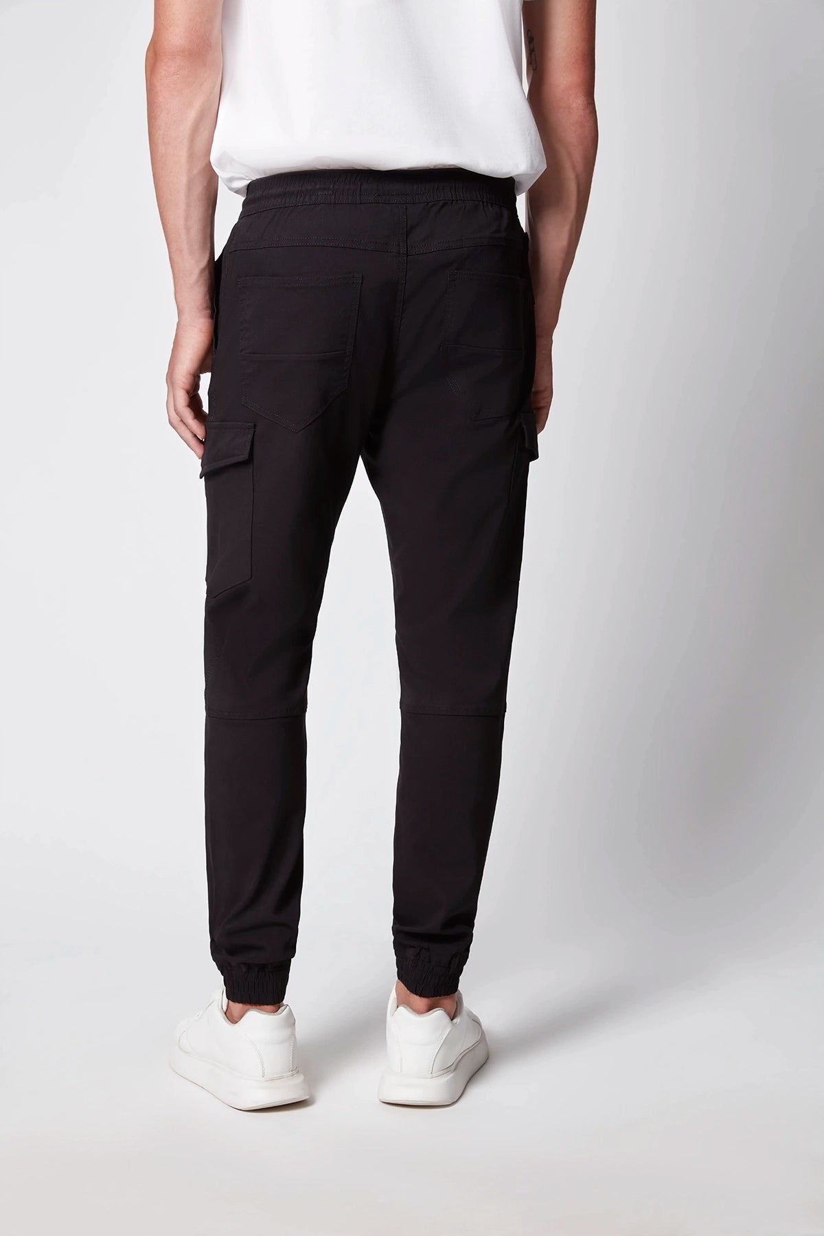 A person wearing a white shirt, PROJEK RAW Men’s Stretch Bungee Cargo Jogger in black, and white sneakers stands facing away from the camera against a plain light background.