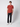 A young man stands against a white background, wearing a PROJEK RAW Mens Short Sleeve Crewneck T-shirt in rust, paired with dark jeans and white sneakers. He faces forward with a neutral expression and a relaxed posture.