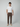 A man poses against a plain light background, wearing a white t-shirt, beige slip-ons, and PROJEK RAW Mens Solid Bungee Fashion Chino Pant in brown with a tailored fit. One hand is in his pocket as he looks at the camera.