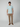 A man stands facing forward against a plain light background, wearing a light blue short-sleeve T-shirt, brown slip-on shoes, and tan PROJEK RAW Mens Solid Bungee Relaxed 5-Pocket Pant with an elasticized waistband. His arms rest by his sides.