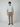 A man with short curly hair and a neutral expression stands against a plain light gray background, wearing PROJEK RAW Mens Solid Bungee Relaxed 5-Pocket Pants and a light blue t-shirt, paired with brown loafers.