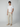 A young man with curly brown hair wears a plain white t-shirt, PROJEK RAW Mens Drawstring Chino Pant in light beige, and beige slip-on shoes. He stands against a gray background, one hand in his pocket, looking at the camera.