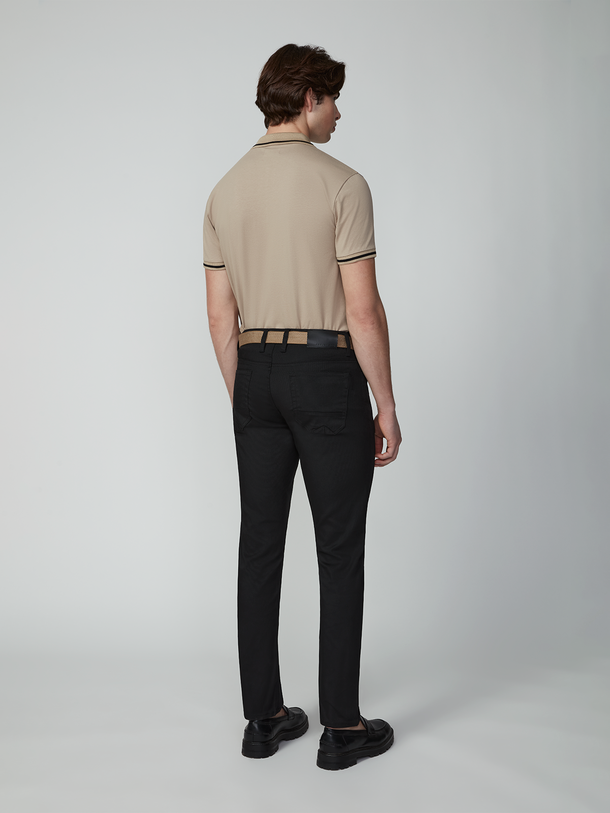 A man faces away from the camera wearing a beige short-sleeve polo, PROJEK RAW MENS 5-POCKET PANT in black, a black belt, and black shoes against a plain light gray background.