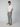 A young man stands against a plain background wearing a white short-sleeve t-shirt, PROJEK RAW MENS 5-POCKET PANT made from stretch fabric, and tan suede sneakers, with his left hand in his pocket.