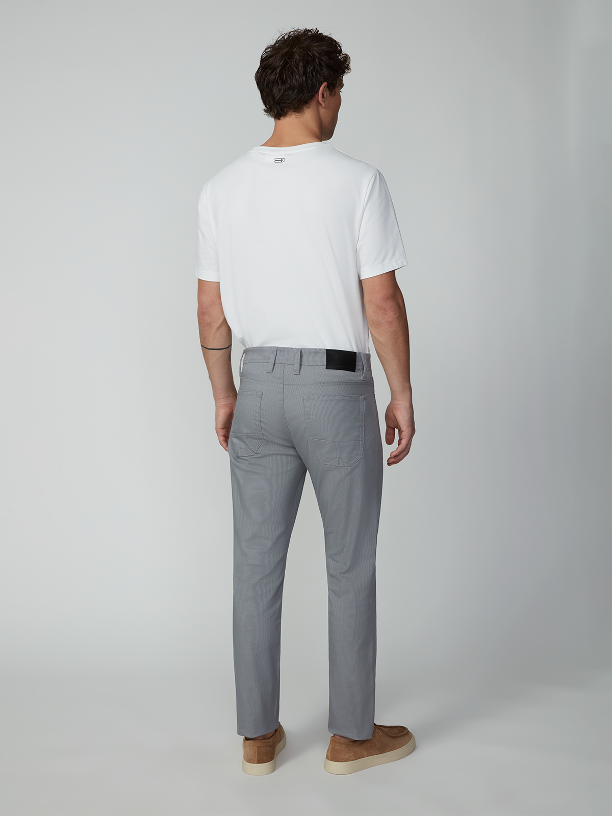 A man faces away from the camera in a white T-shirt and PROJEK RAW MENS 5-POCKET PANT made of stretch fabric, paired with tan shoes, set against a plain light gray background.