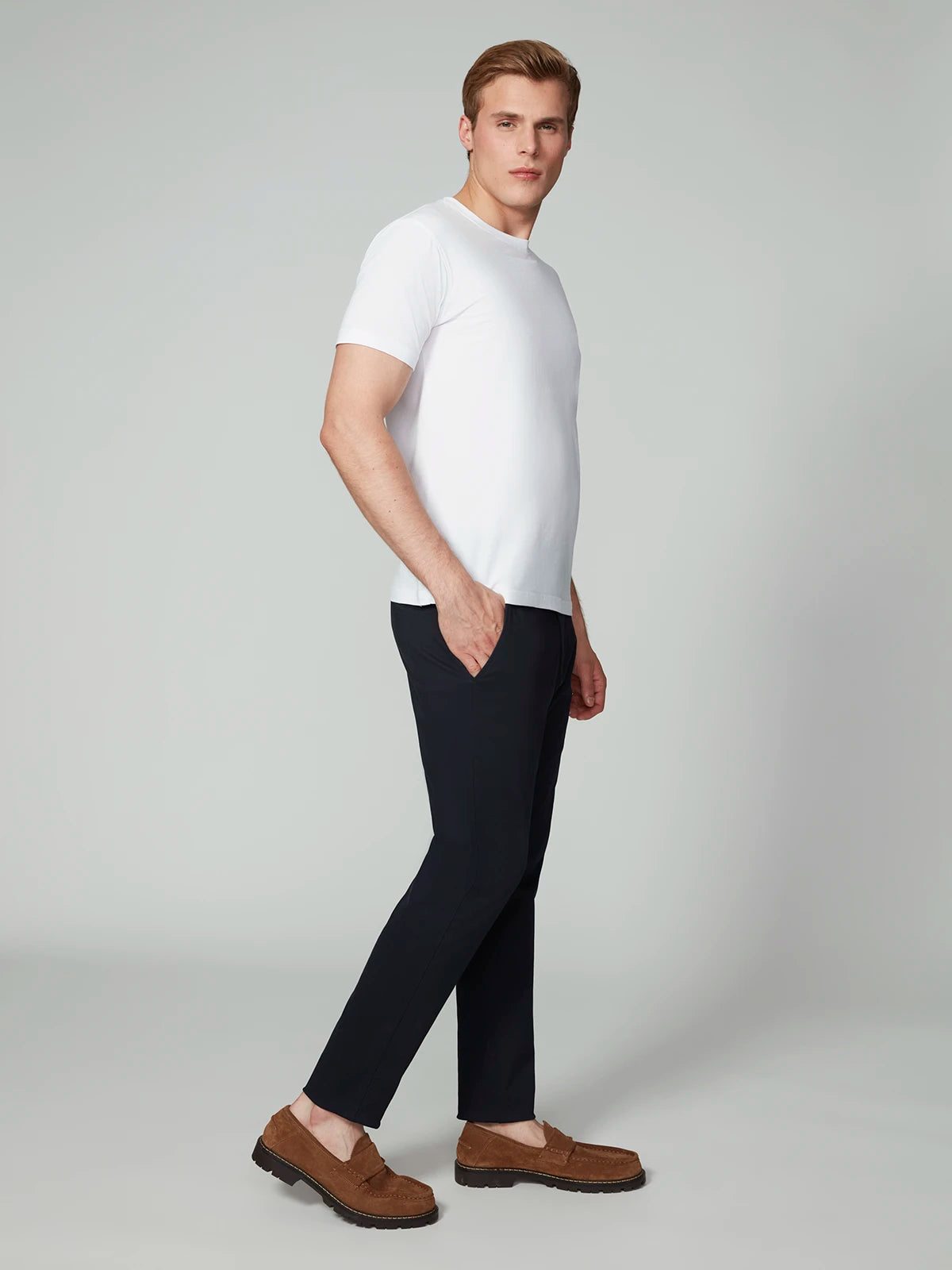 A man stands sideways with his hands in his pockets, wearing a white short-sleeve t-shirt, PROJEK RAW MENS CHINO SIGNATURE WOVEN PANT in black, and brown loafers against a plain light gray background.