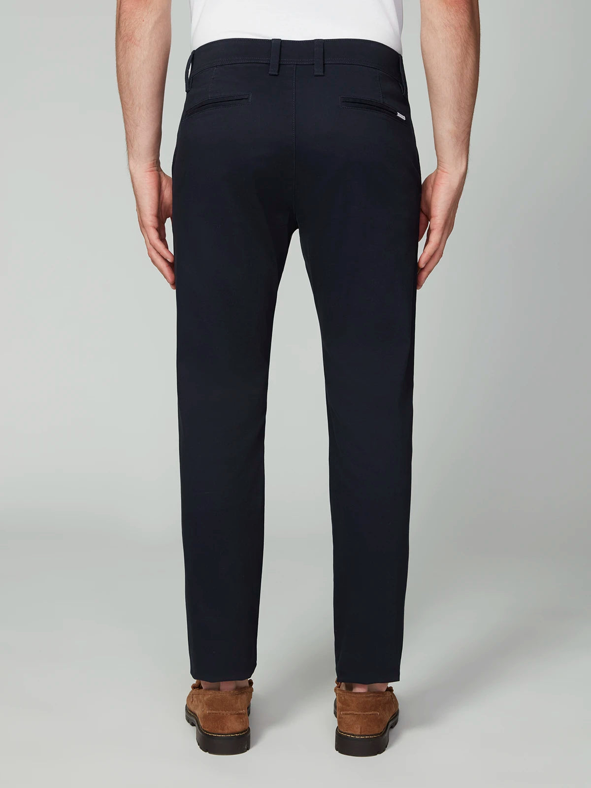 A man faces away wearing PROJEK RAW MENS CHINO SIGNATURE WOVEN PANT in dark navy, styled with a white shirt and brown suede loafers. The tailored pants offer belt loops, buttoned back pockets, and a refined silhouette.