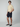 A young man stands against a plain background, wearing a light yellow knit polo shirt, PROJEK RAW Mens 5-Pocket Solid Bungee Shorts in dark navy, and dark slip-on shoes. He faces slightly to the side with a neutral expression.