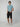A young man smiles, wearing a light blue T-shirt, PROJEK RAW Mens Solid Bungee Capri Cargo Shorts with a 13 inseam and cargo pockets, plus white sneakers. He stands against a plain light gray background with one hand in his pocket.