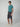A young man stands against a light gray background, wearing a teal short-sleeve t-shirt, PROJEK RAW Mens Solid Bungee Capri Cargo Shorts 13 Inseam with cargo pockets, and beige sneakers. He looks forward neutrally, one hand in his pocket.