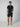 A man poses against a plain background wearing PROJEK RAWs Mens Texture Bungee 5-Pocket Short—black with an elastic waistband—paired with a navy short-sleeve polo with white trim and beige slip-on shoes, one hand in his pocket as he looks at the camera.