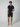 A man stands against a plain background wearing a navy short-sleeve polo shirt, PROJEK RAW Mens Texture Bungee 5-Pocket Shorts with an elastic waistband, and beige slip-on shoes. One hand is in his pocket as he looks slightly to the side.