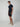 A man with short curly hair and a neutral expression wears a navy polo, PROJEK RAW Mens Texture Bungee 5-Pocket Shorts with an elastic waistband in black, and beige slip-on shoes, standing against a plain background.