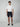A young man stands facing forward against a plain light background, wearing PROJEK RAW Mens Texture Bungee 5-Pocket Short with an elastic waistband, a white t-shirt, and dark slip-on shoes, his posture relaxed and expression neutral.