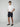 A young man stands against a plain background, wearing PROJEK RAW Mens Texture Bungee 5-Pocket Shorts in navy with an elastic waistband, a white t-shirt, and navy slip-on shoes. One hand is in his pocket as he looks at the camera.