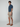 A young man faces sideways in a navy short-sleeve button-up, PROJEK RAW Mens Texture Bungee 5-Pocket Short in light beige, and brown boat shoes. The scene is set against a plain light gray background.