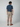 A man, facing away, wears a navy short-sleeve shirt, brown loafers, and PROJEK RAW Mens Texture Bungee 5-Pocket Shorts. The background is plain and light-colored.