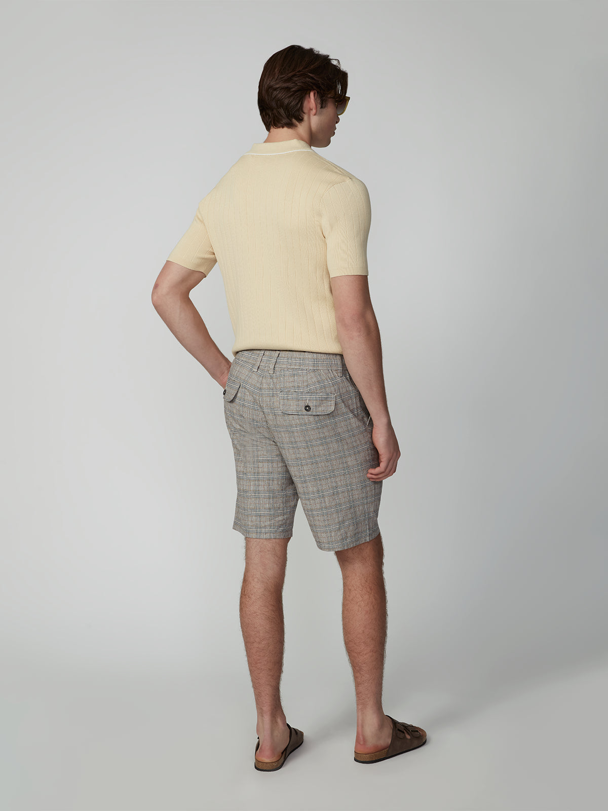 A man faces away, wearing a light beige short-sleeve shirt, PROJEK RAW MENS PLEATED DRAWSTRING CHINO SHORT in gray, brown sandals, and sunglasses against a plain light background—perfect for effortless summer style.
