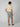 A man faces away, wearing a light beige short-sleeve shirt, PROJEK RAW MENS PLEATED DRAWSTRING CHINO SHORT in gray, brown sandals, and sunglasses against a plain light background—perfect for effortless summer style.