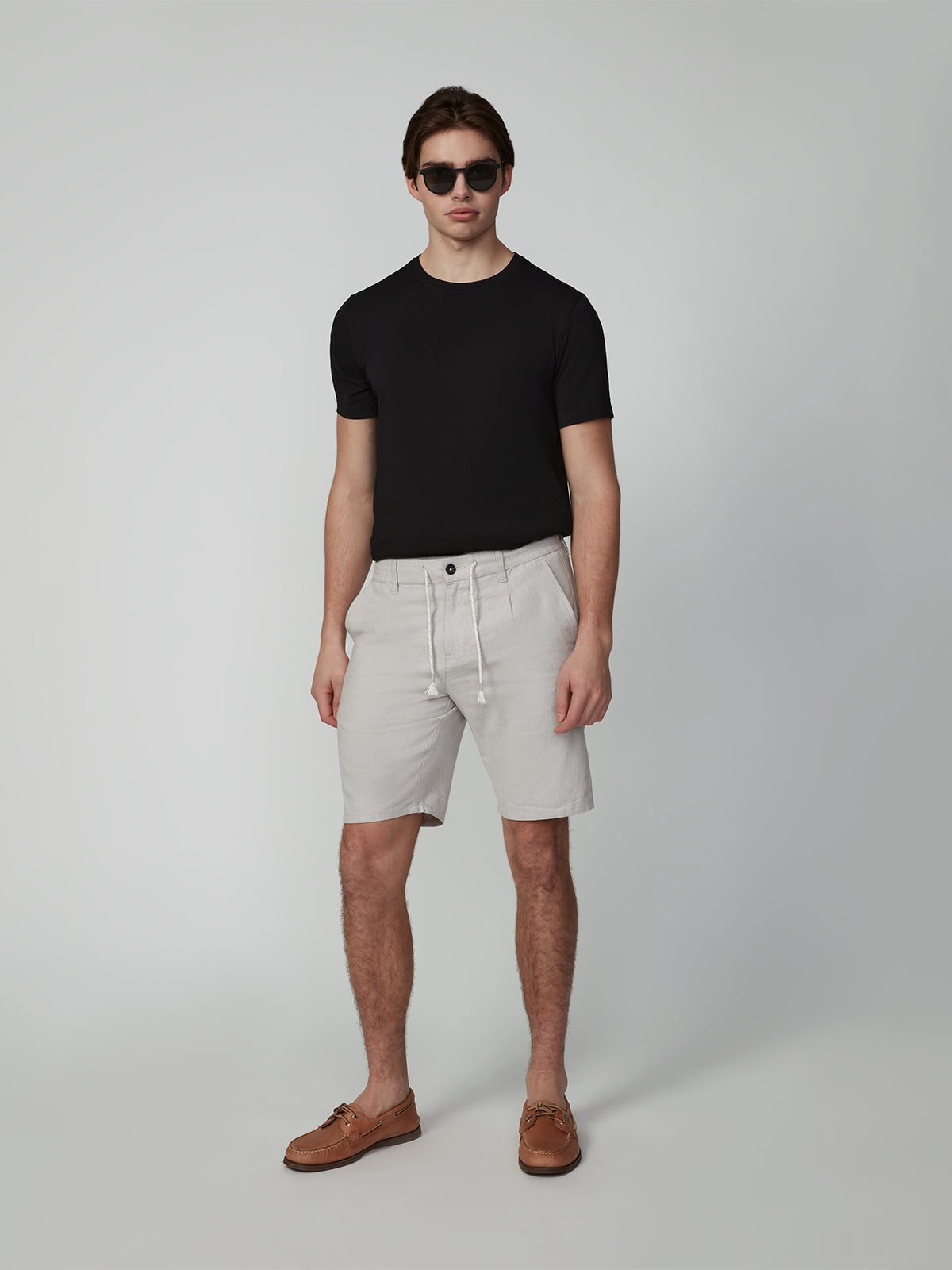 A man in sunglasses, a black t-shirt, PROJEK RAW MENS PLEATED DRAWSTRING CHINO SHORTS, and brown boat shoes stands against a plain light gray background.