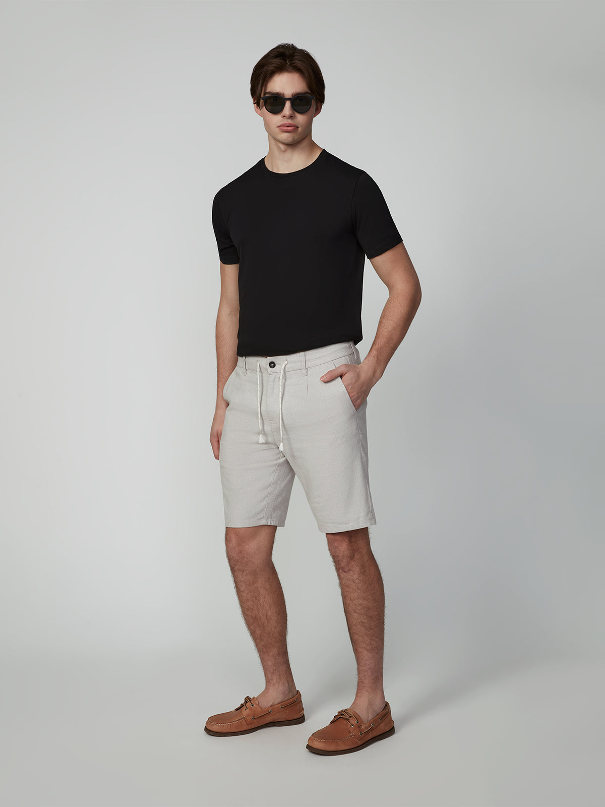 A man in dark sunglasses, a black t-shirt, PROJEK RAW MENS PLEATED DRAWSTRING CHINO SHORTS, and brown boat shoes stands in a neutral studio with his hands in his pockets.