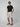 A man in dark sunglasses, a black t-shirt, PROJEK RAW MENS PLEATED DRAWSTRING CHINO SHORTS, and brown boat shoes stands in a neutral studio with his hands in his pockets.
