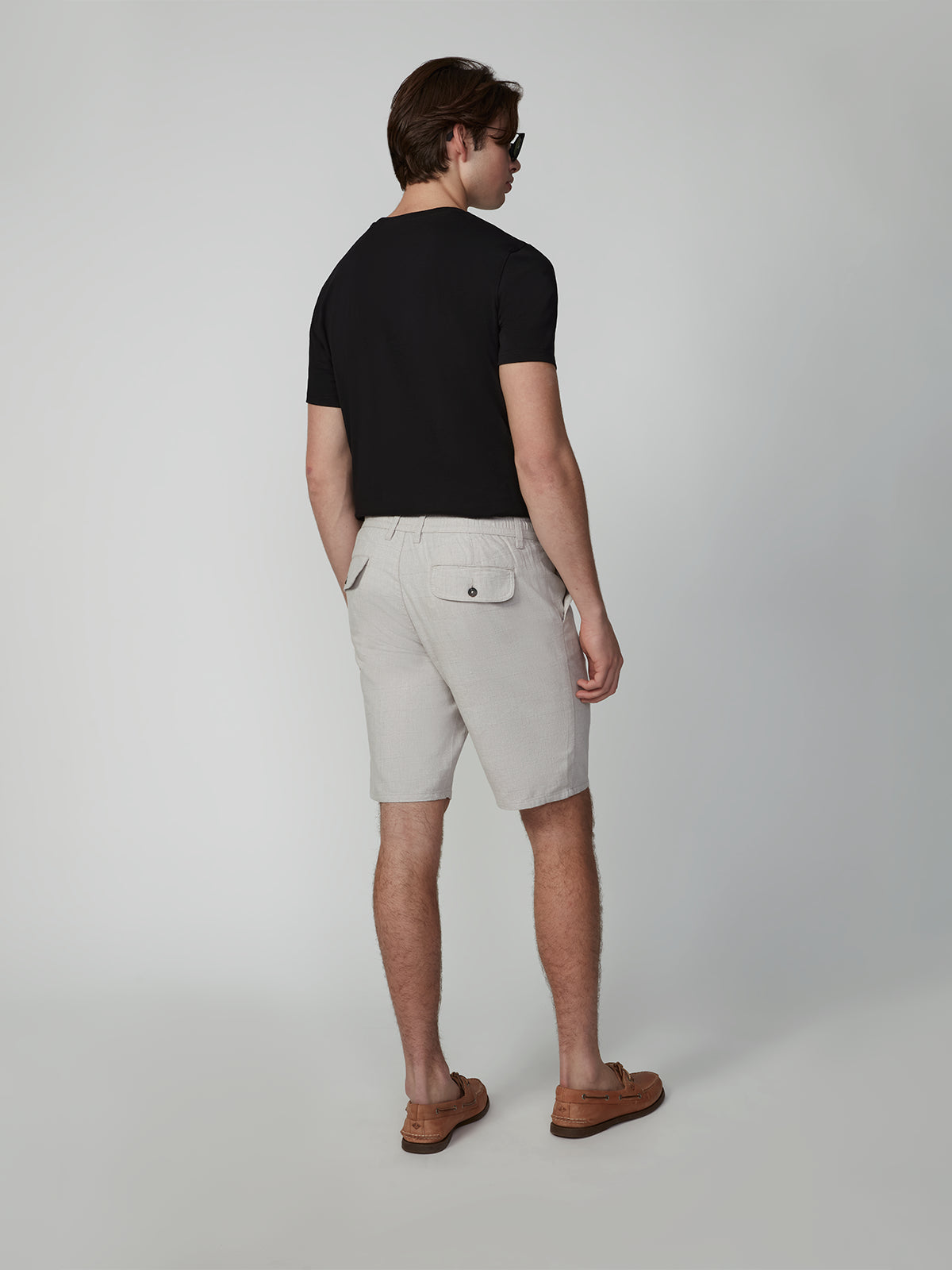 A man with short brown hair and sunglasses wears PROJEK RAW MENS PLEATED DRAWSTRING CHINO SHORTS in light beige, a black t-shirt, and brown loafers with his hands in the back pockets, standing against a plain light background.