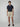 A man with short brown hair and a neutral expression wears PROJEK RAW MENS CHINO SIGNATURE SHORT 8, paired with a navy short-sleeve button-up shirt, belt, and brown loafers against a plain background, showcasing everyday comfort and tailored fit.