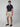 A young man stands against a plain background wearing PROJEK RAW MENS CHINO SIGNATURE SHORT 8, paired with a navy short-sleeve shirt, belt, and brown loafers. He gazes ahead neutrally, showcasing relaxed everyday style.