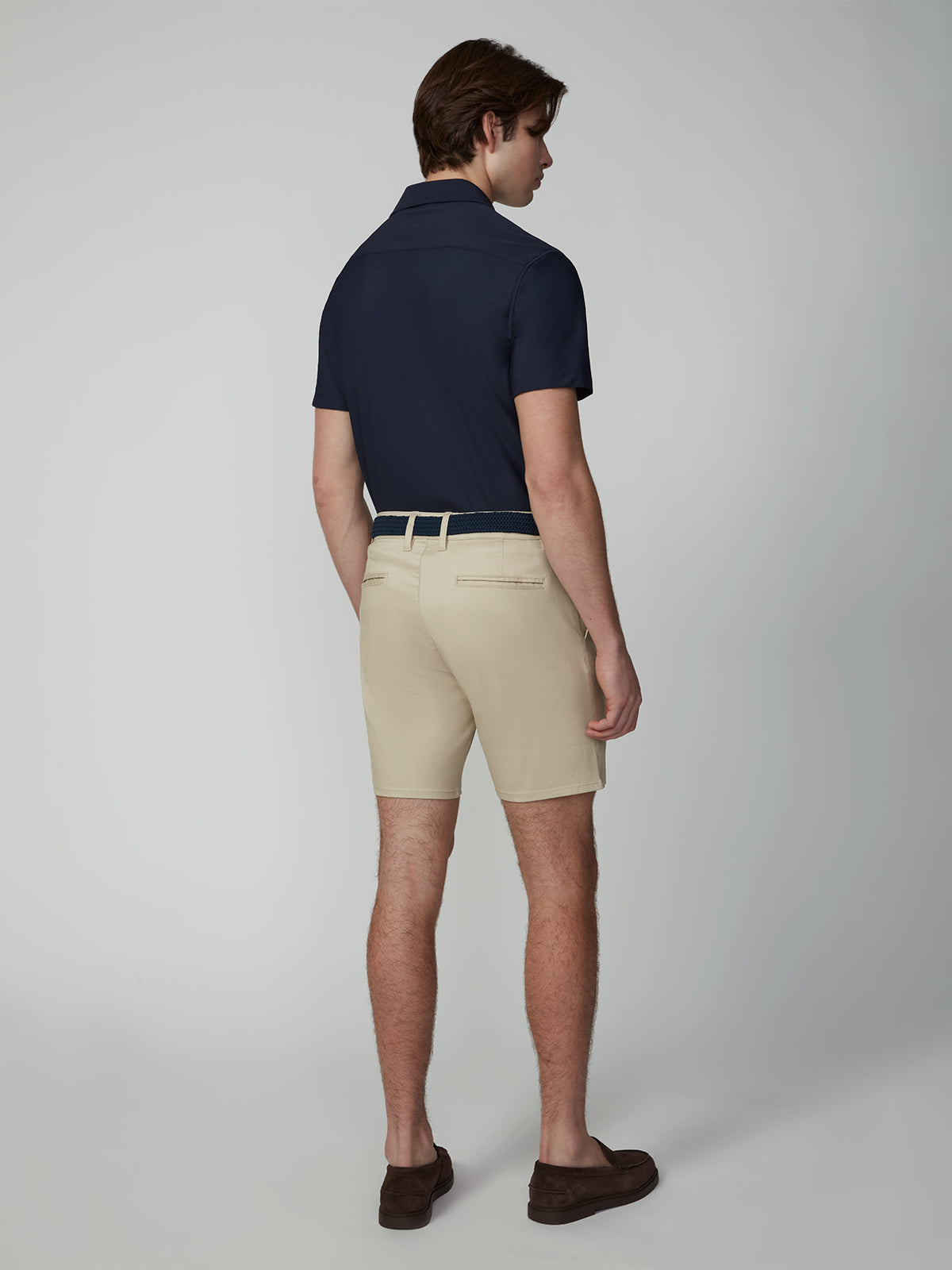 A man faces away, wearing a navy short-sleeve polo, PROJEK RAW MENS CHINO SIGNATURE SHORT 8 in beige with a belt, and brown loafers. The background is plain and light-colored.