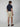 A man faces away, wearing a navy short-sleeve polo, PROJEK RAW MENS CHINO SIGNATURE SHORT 8 in beige with a belt, and brown loafers. The background is plain and light-colored.