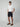 A young man stands against a plain light background, wearing a white polo, brown loafers, and the PROJEK RAW MENS CHINO SIGNATURE SHORT 8 in black with a tailored fit. One hand is in his pocket as he looks directly at the camera with a neutral expression.