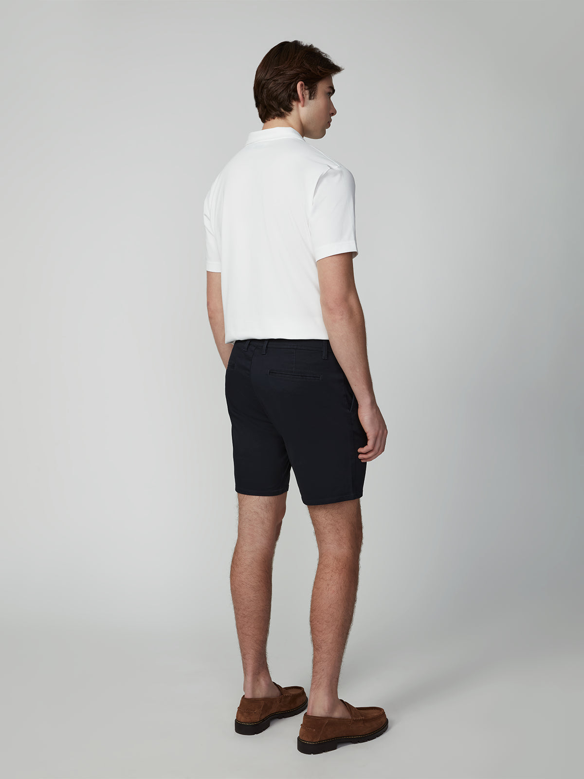 A man faces away from the camera wearing a white short-sleeve polo, PROJEK RAW MENS CHINO SIGNATURE SHORT 8 in dark navy, and brown loafers against a plain light gray background.
