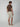 A man faces away from the camera wearing a brown short-sleeve shirt, PROJEK RAW MENS DRAWSTRING CHINO SHORT in light beige, and beige slip-on shoes against a plain light background.