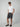 A man in sunglasses, a white t-shirt, PROJEK RAW MENS DRAWSTRING CHINO SHORT, and black slip-on shoes stands with hands in pockets against a plain light gray background.