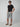 A man in sunglasses, a black t-shirt, PROJEK RAW MENS CARGO LINEN CAPRI SHORTS, and brown sandals stands with one hand in his pocket against a plain light gray background.