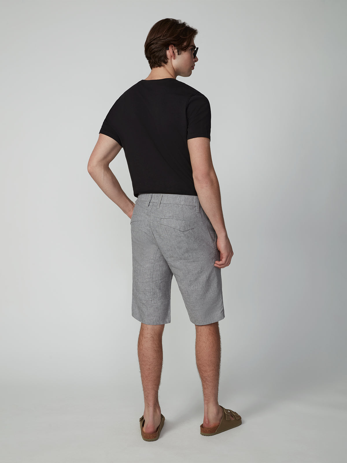 A man faces away against a plain light background, wearing PROJEK RAW MENS CARGO LINEN CAPRI SHORT in light gray, a black t-shirt, brown sandals, and sunglasses. His right hand is on his hip.