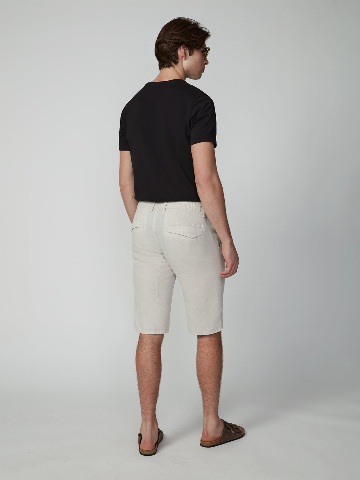 A man faces away from the camera wearing a black t-shirt, brown sandals, and PROJEK RAW MENS CARGO LINEN CAPRI SHORT with multiple pockets. The breathable linen shorts offer all-day comfort against a plain light gray background.