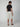 A man faces away from the camera wearing a black t-shirt, brown sandals, and PROJEK RAW MENS CARGO LINEN CAPRI SHORT with multiple pockets. The breathable linen shorts offer all-day comfort against a plain light gray background.