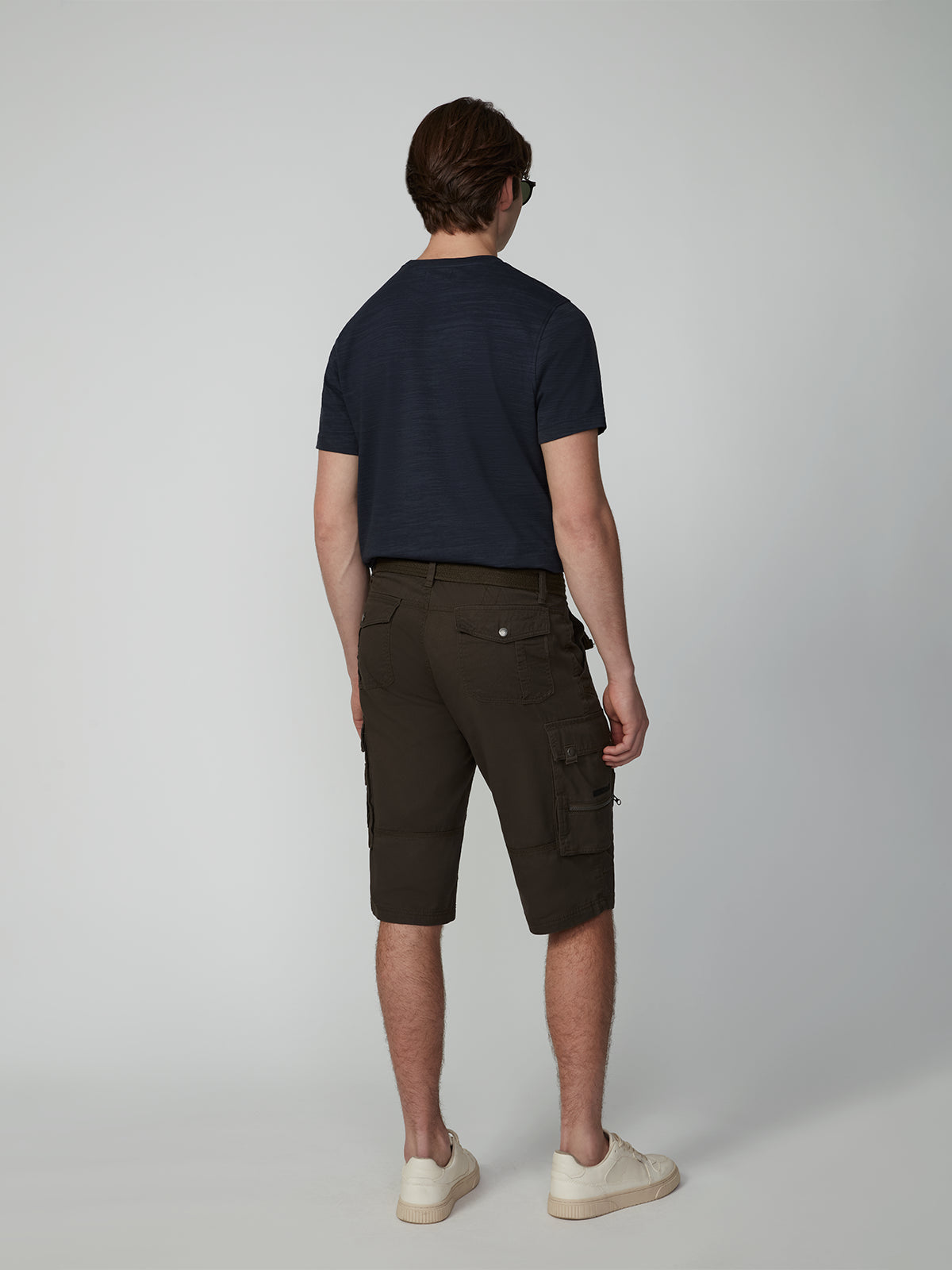 A man, seen from behind, wears PROJEK RAW MENS CARGO CAPRI SHORTS with a navy t-shirt, white sneakers, and sunglasses against a plain light gray background.