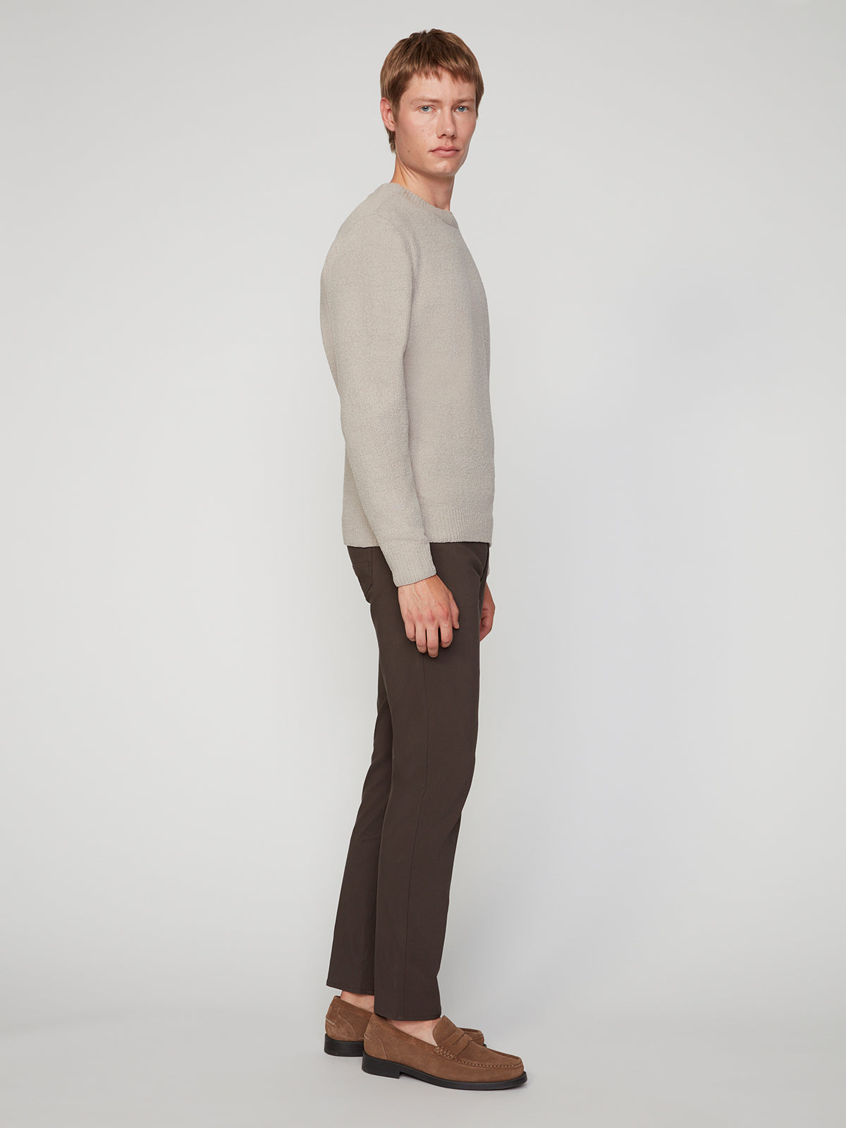 A man stands sideways against a white background, wearing a light beige sweater, brown loafers, and PROJEK RAW MENS 5-POCKET SOLID BUNGEE PANT in dark brown. He looks forward with a neutral expression and his hands by his sides.