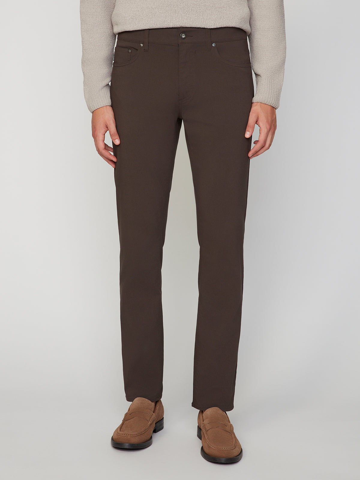 A person is shown from shoulders to feet wearing PROJEK RAW MENS 5-POCKET SOLID BUNGEE PANT in dark brown, paired with a light beige sweater and brown loafers, standing against a plain white background.