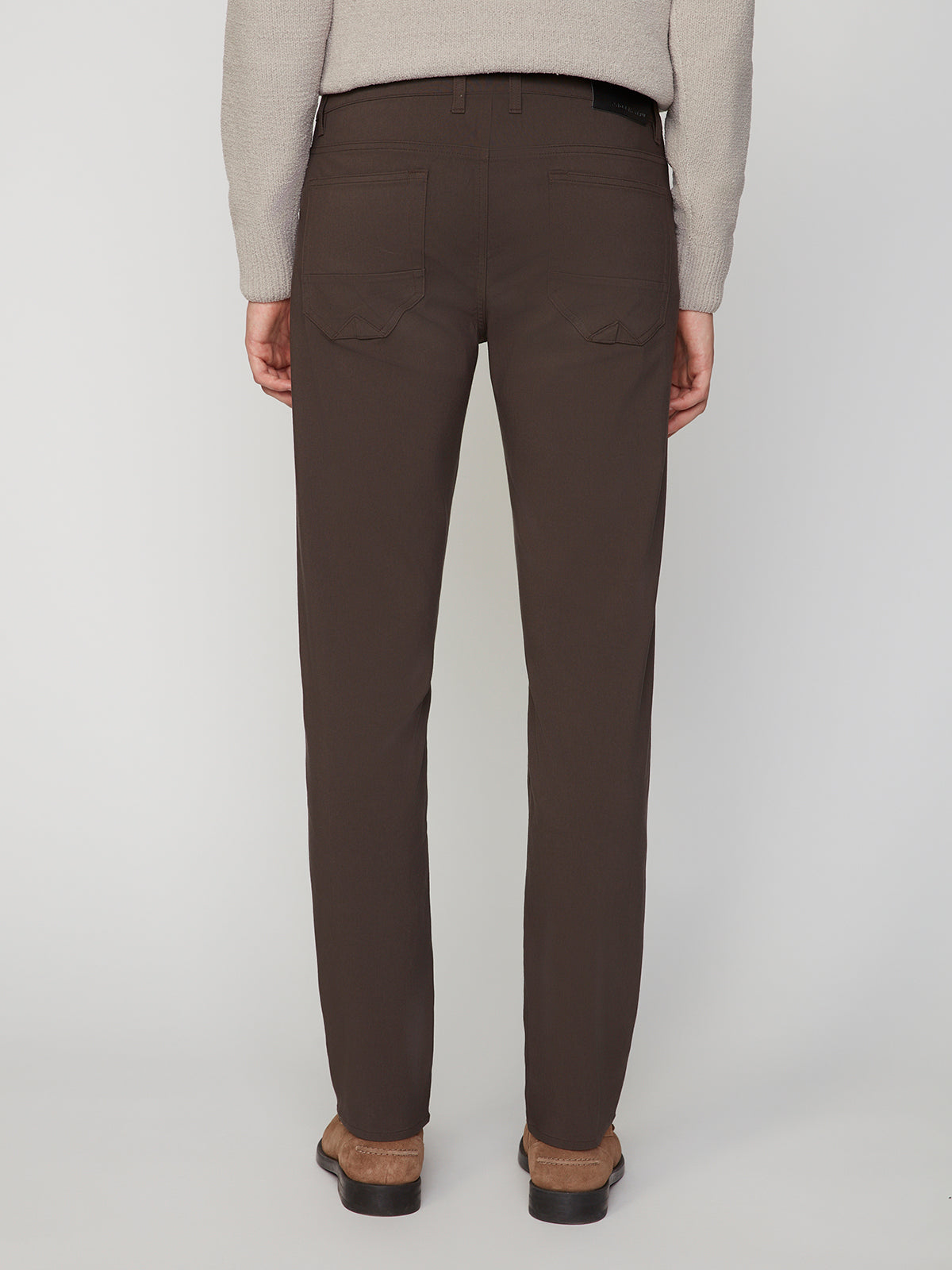 A person faces away, wearing PROJEK RAW MENS 5-POCKET SOLID BUNGEE PANT in dark brown—classic fit with flap back pockets. Styled with a light beige sweater and brown shoes, the outfit pops against a simple, light background.