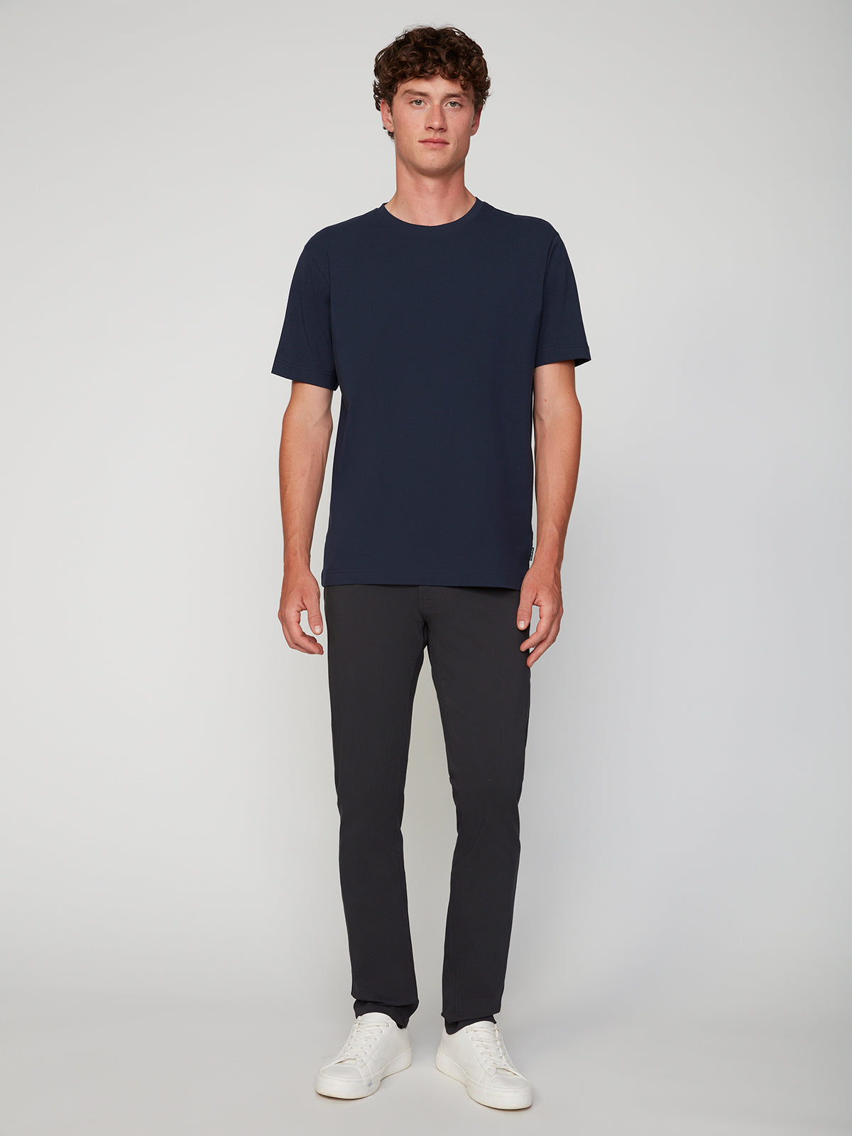 A person with short curly brown hair stands facing forward against a light gray background, wearing a plain navy t-shirt, white sneakers, and PROJEK RAW MENS 5-POCKET SOLID BUNGEE PANT with an elastic waistband.