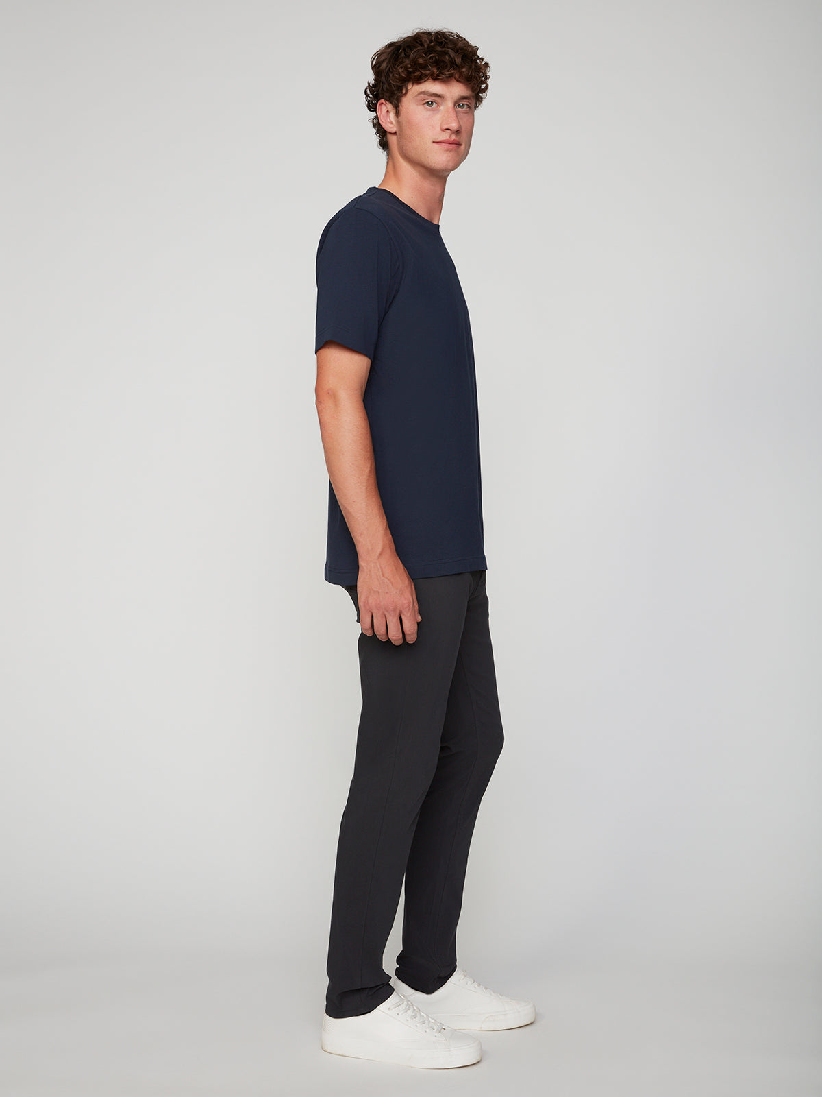 A young man with curly hair stands in profile against a plain light background, wearing a navy blue t-shirt, PROJEK RAW MENS 5-POCKET SOLID BUNGEE PANT with an elastic waistband, and white sneakers, looking straight ahead neutrally.