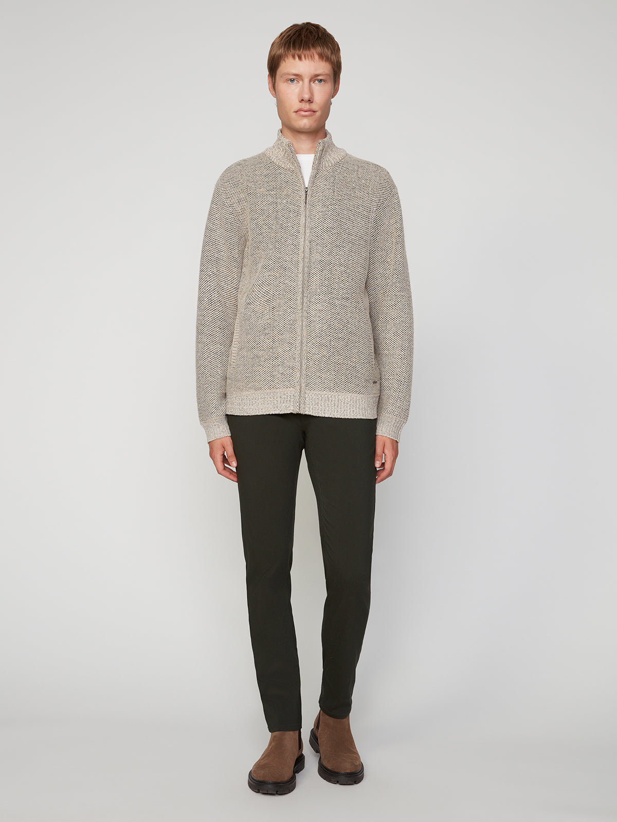 A person stands against a plain light background wearing PROJEK RAW MENS 5-POCKET SOLID BUNGEE PANT in dark green, paired with a beige zip-up cardigan and brown shoes. They face forward with a neutral expression and relaxed posture.