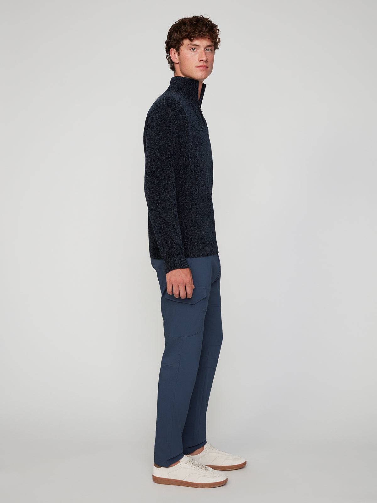 A young man stands sideways against a light gray background, wearing PROJEK RAW MENS SOLID BUNGEE CARGO PANT in blue, a navy zip-up sweater, and white sneakers.