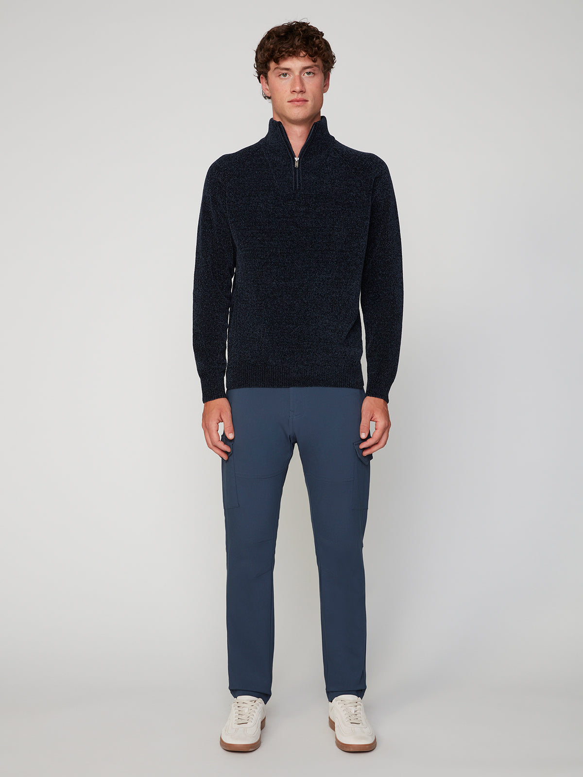 A man with short curly hair stands facing forward in a minimalist streetwear look, wearing PROJEK RAW MENS SOLID BUNGEE CARGO PANT, a dark half-zip sweater, and white sneakers against a plain background.