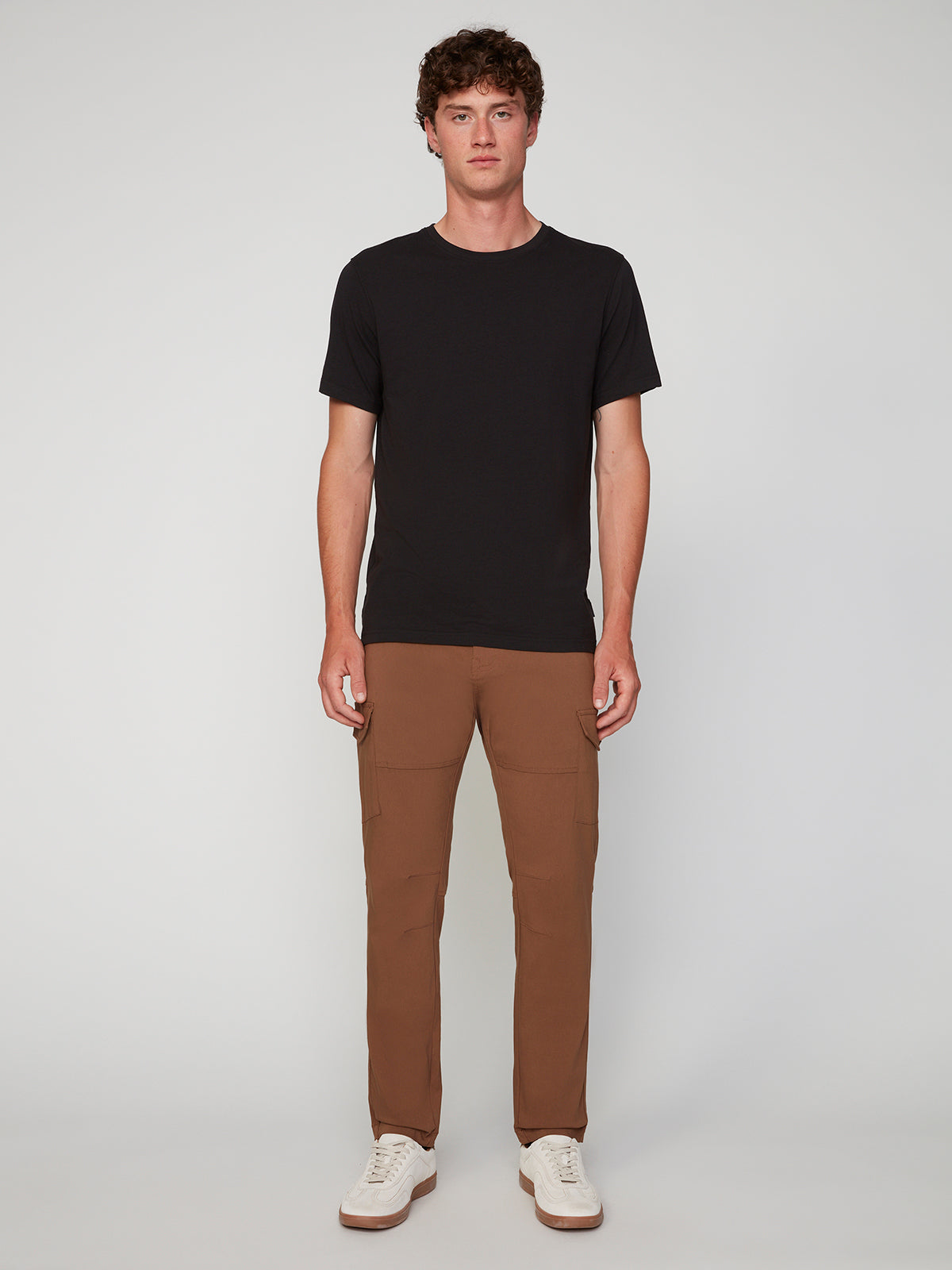 A young man with short curly hair wears a plain black t-shirt, white sneakers, and PROJEK RAW MENS SOLID BUNGEE CARGO PANT with an elasticized waistband, standing facing forward against a light gray background.