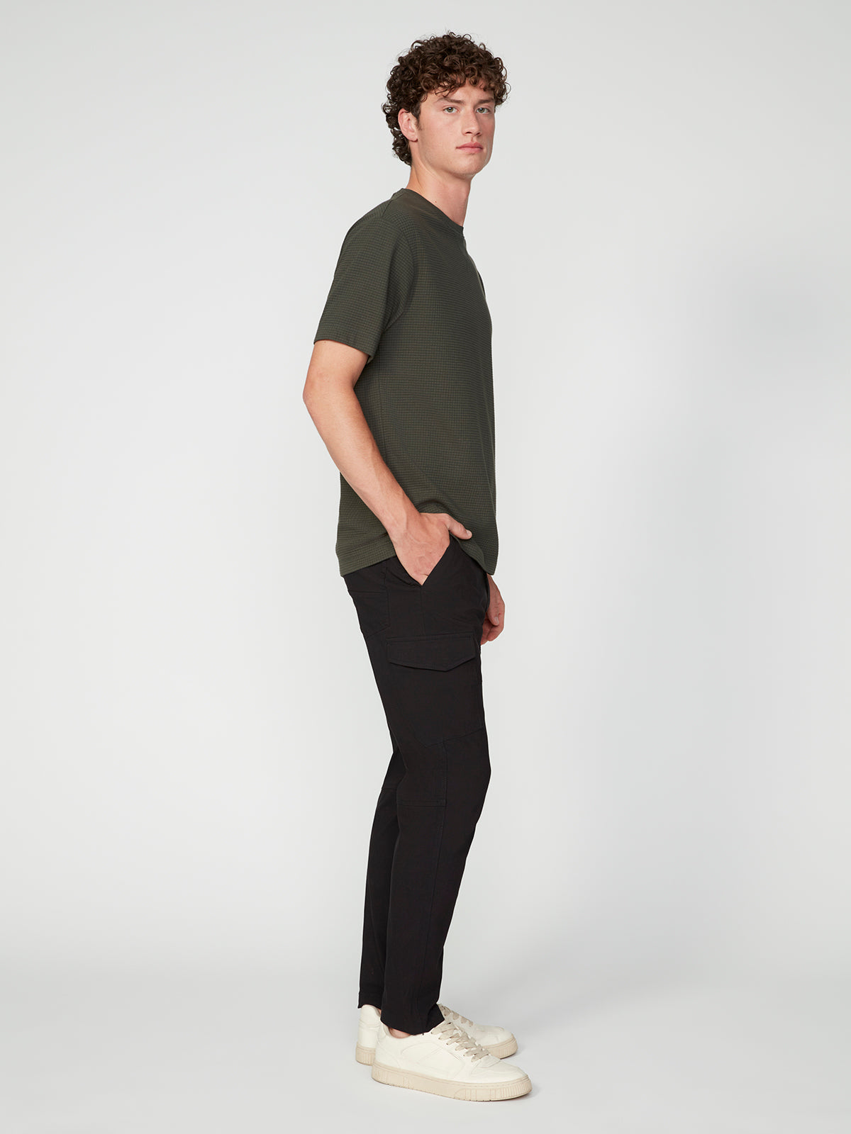 A young man with curly hair stands in profile against a plain background, wearing streetwear: a dark green t-shirt, PROJEK RAW MENS SOLID BUNGEE CARGO PANT with cargo pockets, and white sneakers, one hand casually in his pocket.